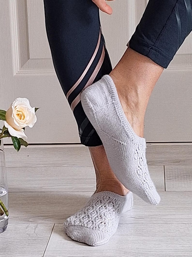 May include: Pale grey knitted no-show socks with an intricate openwork design. The socks are worn with black athletic leggings featuring rose gold stripes. A white rose is in a glass vase.