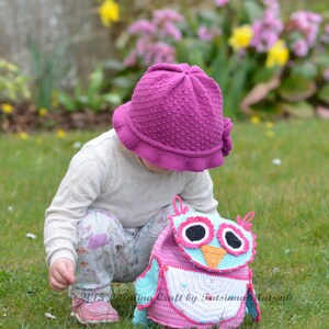 May include: A young child wearing a pink crocheted hat looks down at a pink and blue crocheted owl backpack. The backpack has large black eyes and an orange beak. The child is standing on a grassy lawn with yellow flowers in the background.