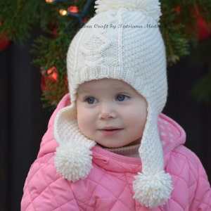 May include: A young child wearing a white knitted hat with a pom-pom on top and a pink quilted jacket. The hat has ear flaps and a white pom-pom on each flap. The child is looking at the camera.