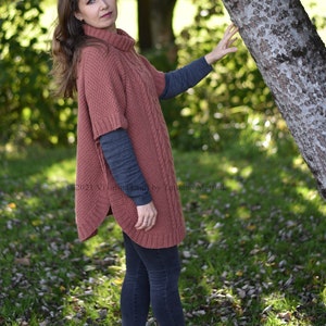 K&ouml;nnte beinhalten: Eine Frau tr&auml;gt einen rostigen Strickponcho mit Zopfmuster, dunkelblaue Jeans und rote Schuhe. Sie steht in einem grasbewachsenen Bereich mit heruntergefallenen Bl&auml;ttern.