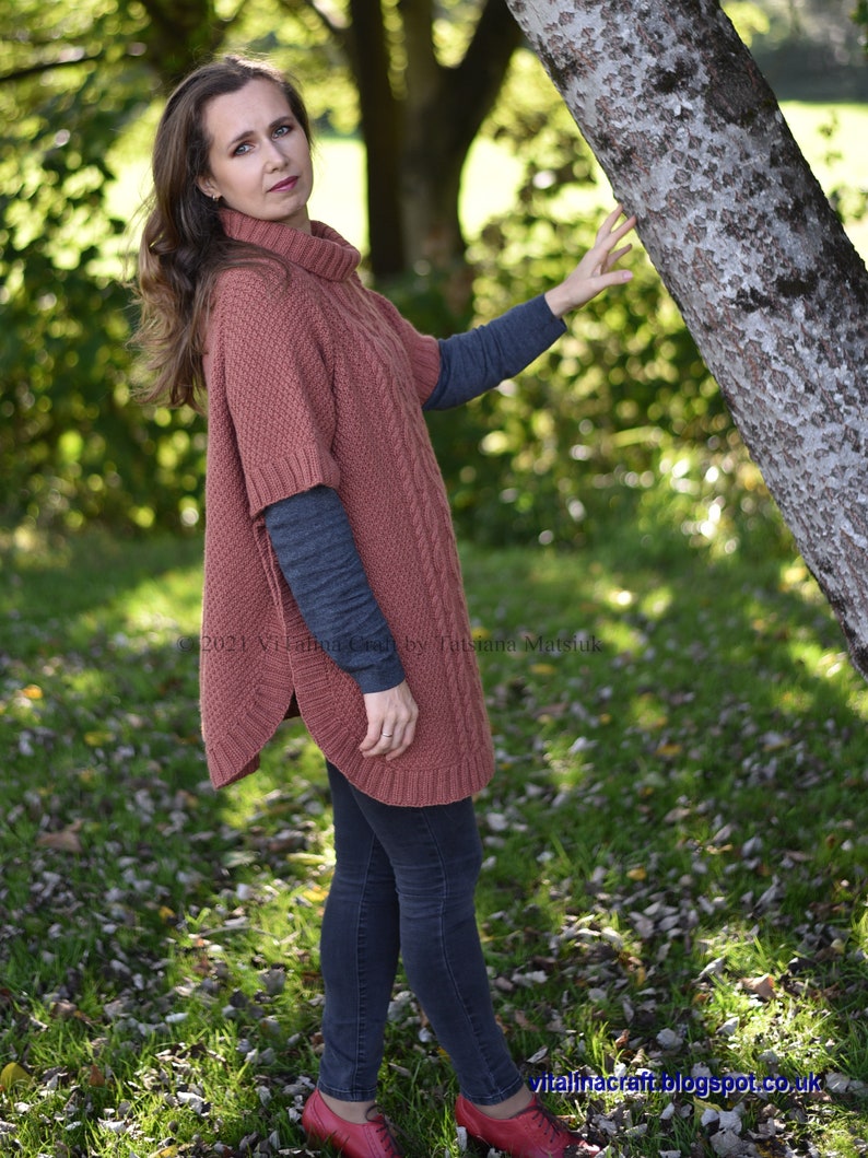K&ouml;nnte beinhalten: Eine Frau tr&auml;gt einen rostigen Strickponcho mit Zopfmuster, dunkelblaue Jeans und rote Schuhe. Sie steht in einem grasbewachsenen Bereich mit heruntergefallenen Bl&auml;ttern.
