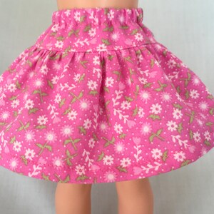 Puede incluir: Una falda rosa con estampado floral con flores blancas y hojas verdes. La falda tiene una cintura fruncida y un largo que llega justo por encima de las rodillas.