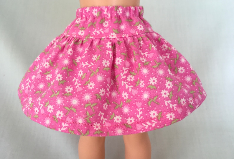 Puede incluir: Una falda rosa con estampado floral con flores blancas y hojas verdes. La falda tiene una cintura fruncida y un largo que llega justo por encima de las rodillas.