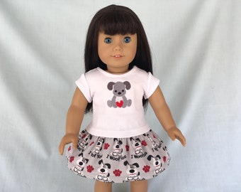 May include: A white t-shirt with a gray dog wearing a red heart on it. The doll is wearing a gray skirt with a pattern of gray dogs and the text "I woof you".