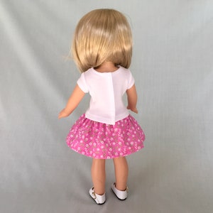 Puede incluir: Una mu&ntilde;eca con una camisa blanca de manga corta y una falda rosa con estampado floral. La mu&ntilde;eca tiene el pelo rubio y est&aacute; de pie sobre un fondo gris claro.