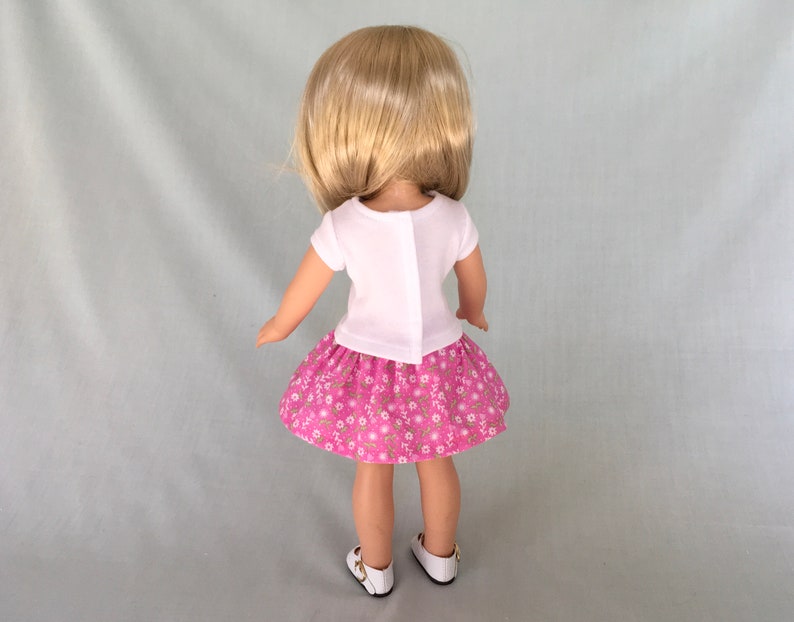 Puede incluir: Una mu&ntilde;eca con una camisa blanca de manga corta y una falda rosa con estampado floral. La mu&ntilde;eca tiene el pelo rubio y est&aacute; de pie sobre un fondo gris claro.
