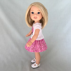 Puede incluir: Una mu&ntilde;eca con una camiseta blanca con un dise&ntilde;o de mariposa rosa y una falda rosa con flores. La mu&ntilde;eca tiene el pelo rubio y los ojos azules.