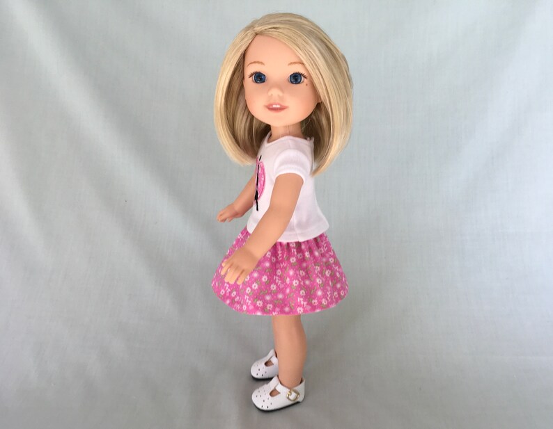Puede incluir: Una mu&ntilde;eca con una camiseta blanca con un dise&ntilde;o de mariposa rosa y una falda rosa con flores. La mu&ntilde;eca tiene el pelo rubio y los ojos azules.