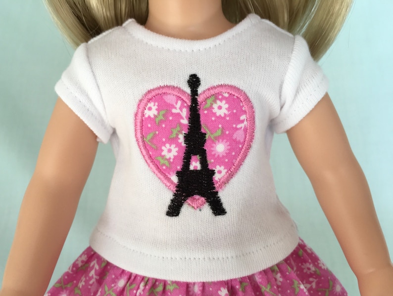 Puede incluir: Camiseta blanca con un parche rosa en forma de coraz&oacute;n que presenta una silueta negra de la Torre Eiffel. El parche en forma de coraz&oacute;n tiene un patr&oacute;n floral.