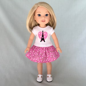 Puede incluir: Una mu&ntilde;eca con una camiseta blanca con un coraz&oacute;n rosa y un dise&ntilde;o de la Torre Eiffel negra. La mu&ntilde;eca tambi&eacute;n lleva una falda rosa con flores blancas y zapatos blancos.