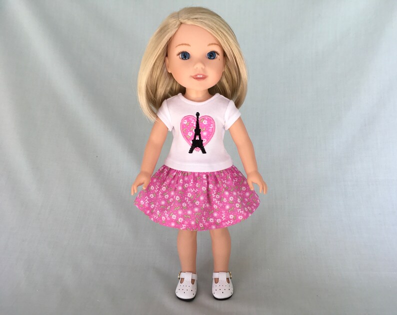 Puede incluir: Una mu&ntilde;eca con una camiseta blanca con un coraz&oacute;n rosa y un dise&ntilde;o de la Torre Eiffel negra. La mu&ntilde;eca tambi&eacute;n lleva una falda rosa con flores blancas y zapatos blancos.