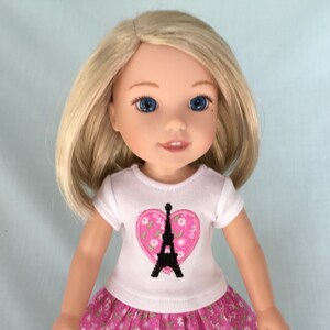 Puede incluir: Una mu&ntilde;eca con cabello rubio que lleva una camiseta blanca con un coraz&oacute;n rosa y un dise&ntilde;o de la Torre Eiffel negra. La mu&ntilde;eca tambi&eacute;n lleva una falda floral rosa y blanca.