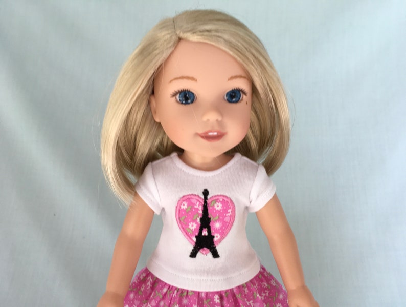Puede incluir: Una mu&ntilde;eca con cabello rubio que lleva una camiseta blanca con un coraz&oacute;n rosa y un dise&ntilde;o de la Torre Eiffel negra. La mu&ntilde;eca tambi&eacute;n lleva una falda floral rosa y blanca.