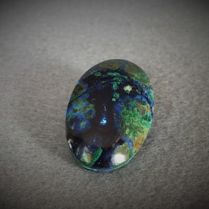 Könnte beinhalten: Ovaler Cabochon-Edelstein mit einem dunkelblauen, grünen und braunen Muster. Der Stein hat eine glatte, polierte Oberfläche.