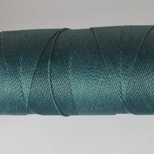 Può includere: Una bobina di filo di nylon color verde acqua. Il filo è avvolto strettamente attorno a un'anima nera, creando una forma cilindrica. Il filo ha una finitura leggermente lucida e un aspetto strutturato, adatto a vari progetti di artigianato.