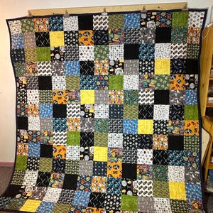 May include: A handmade quilt with a patchwork design. The quilt features a variety of fabrics in squares, including black, yellow, green, and blue. Some squares have character prints. The quilt is bordered with a dark blue fabric.