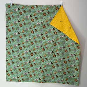 May include: A reversible baby blanket with a jungle animal print on a teal background. The print features lions, elephants, and monkeys. The reverse side is solid yellow. The blanket is hanging on a white wall.