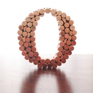 Könnte beinhalten: Ein großer Buchstabe O aus Weinkorken. Die Korken sind in einem Kreis angeordnet, wobei die Enden der Korken nach außen zeigen. Der Buchstabe steht auf einer Holzoberfläche.