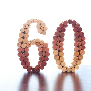 May include: The number 60 is formed using wine corks. The corks are arranged in a gradient pattern, with the darker corks at the bottom and the lighter corks at the top.