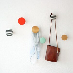 Könnte beinhalten: Eine weiße Wand mit einer braunen Ledertasche, die an Haken hängt. Die Haken sind aus Holz und haben verschiedene Muster, darunter geometrische und einfarbige. Die Tasche hängt an einem Haken mit einem schwarz-weißen geometrischen Muster.