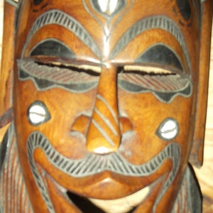 Puede incluir: Una máscara de madera con un rostro tallado, detalles en blanco y negro y acentos blancos alrededor de los ojos. La máscara tiene una boca ancha y una barba tallada.