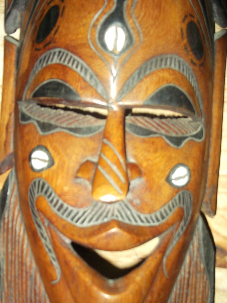 Puede incluir: M&aacute;scara de madera tallada con una cara sonriente. La m&aacute;scara es principalmente marr&oacute;n con detalles en negro y blanco. Los ojos est&aacute;n abiertos y la boca es ancha. Est&eacute;tica tribal o cultural.