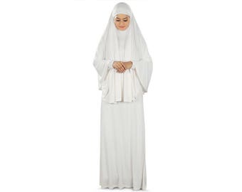 May include: A white modest dress ensemble. The outfit includes a long-sleeved top with a draped overlay, a matching head covering, and a floor-length skirt. The fabric appears soft and flowing, creating a modest and elegant look.