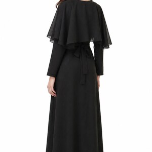 May include: A long black dress with a matching cape. The dress has long sleeves and a flowing skirt. The cape is a sheer, layered design that drapes over the shoulders. A black tie belt cinches the waist. The dress is suitable for formal events.