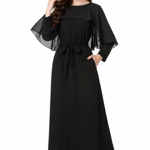 May include: A long, black dress with a round neckline and long sleeves. The dress features a cape-like overlay and a tie belt at the waist. Pockets are visible on the sides. The dress is made of a flowing fabric.