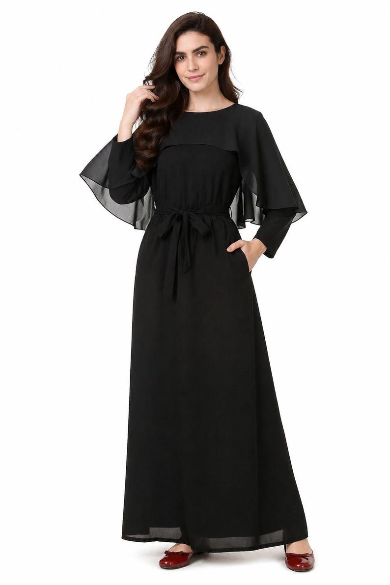 May include: A long, black dress with a round neckline and long sleeves. The dress features a cape-like overlay and a tie belt at the waist. Pockets are visible on the sides. The dress is made of a flowing fabric.