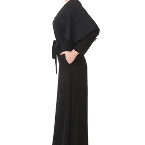 May include: A black jumpsuit with a cape-like overlay and a matching belt. The jumpsuit features long sleeves and wide-leg pants. The model is wearing red flats. The outfit is a sleek and elegant design.