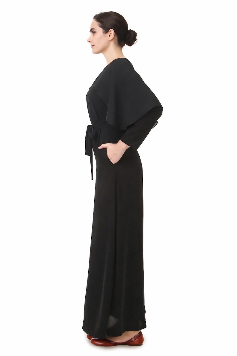 May include: A black jumpsuit with a cape-like overlay and a matching belt. The jumpsuit features long sleeves and wide-leg pants. The model is wearing red flats. The outfit is a sleek and elegant design.