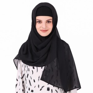 May include: A woman wearing a black hijab and a black underscarf. The hijab is draped over the head and shoulders, with a flowing design. The woman is wearing a white and black patterned top.