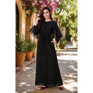 May include: A black maxi dress with a round neckline and long, sheer sleeves. The dress features a waist tie and a flowing skirt. The model is wearing red flats. The dress is suitable for formal occasions.