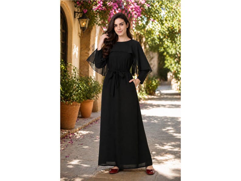 May include: A black maxi dress with a round neckline and long, sheer sleeves. The dress features a waist tie and a flowing skirt. The model is wearing red flats. The dress is suitable for formal occasions.