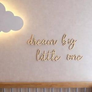 May include: A white cloud-shaped wall light with a warm glow, hanging above a crib. The phrase "dream big little one" is on the wall. The crib contains stuffed animals and a patterned blanket. A nursery decor theme.
