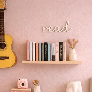 May include: A bedroom with a soft pink colour scheme. A wooden guitar hangs on the wall, next to a shelf of books and a decorative "read" sign. A white cube shelf holds books and storage bins. A bed with a pink pillow and blanket is on the right.