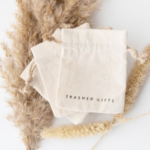 May include: Three natural-colored cotton drawstring bags stacked on top of each other. The top bag has the text "Trashed Gifts" printed in black on the front.