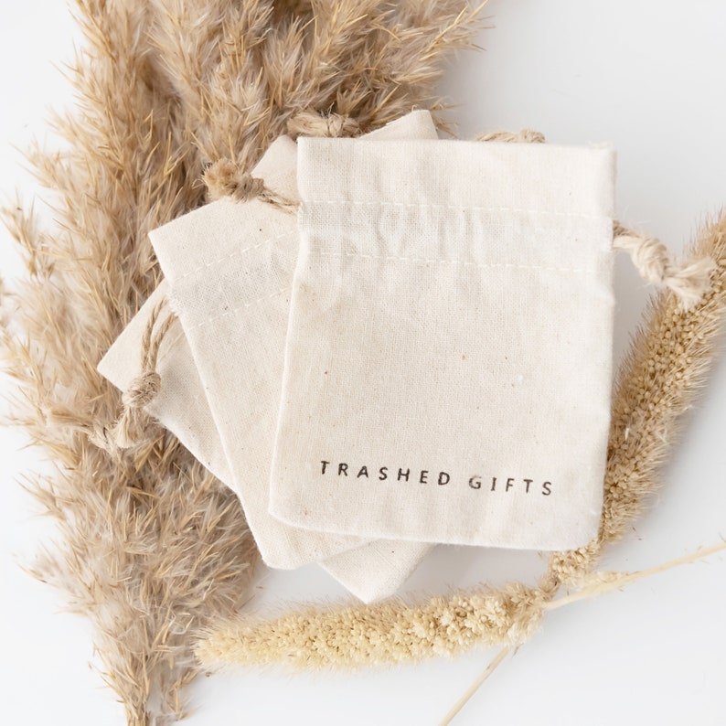 May include: Three natural-colored cotton drawstring bags stacked on top of each other. The top bag has the text "Trashed Gifts" printed in black on the front.