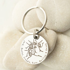 May include: Silver keychain with a round tag featuring a design of five fists clasped together. The tag is engraved with the names "Jack, Daniel, Freddie, Rose, Matilda & Daddy".