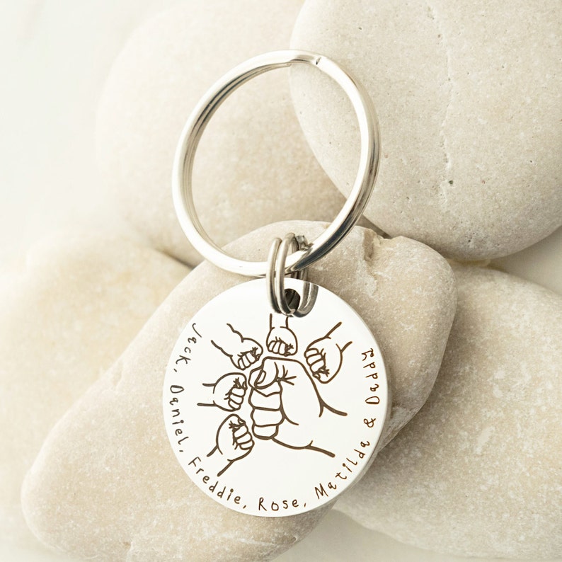 May include: Silver keychain with a round tag featuring a design of five fists clasped together. The tag is engraved with the names "Jack, Daniel, Freddie, Rose, Matilda & Daddy".