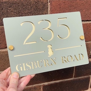 以下が含まれることがあります： 薄緑色の長方形の家の看板。金色の数字「235」が切り抜かれています。犬のシルエットと「GISBURN ROAD」の文字も金色で表示されています。2つの金色のネジで固定されています。メートル法での測定。