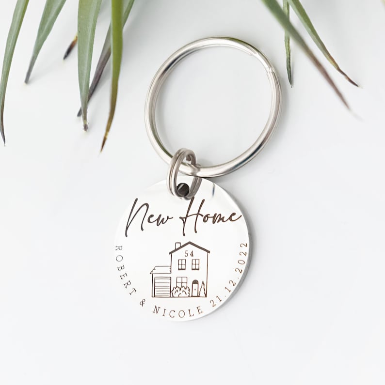 May include: Silver keychain with a round tag engraved with "New Home", a small house illustration, "Robert & Nicole", and the date "21.12.2022".