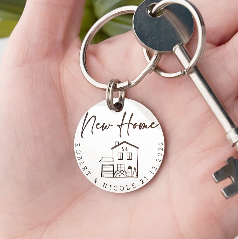 May include: Silver keychain with a round tag that says "New Home" with a house illustration, the names "Robert & Nicole", and the date "21.12.2022".