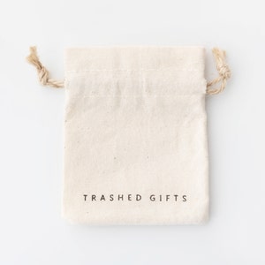 Pode incluir: Uma bolsa de algod&atilde;o branco com cord&atilde;o de ajuste, com o texto "TRASHED GIFTS" impresso na frente.