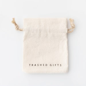 May include: A white cotton drawstring bag with the text "Trashed Gifts" printed on the front.