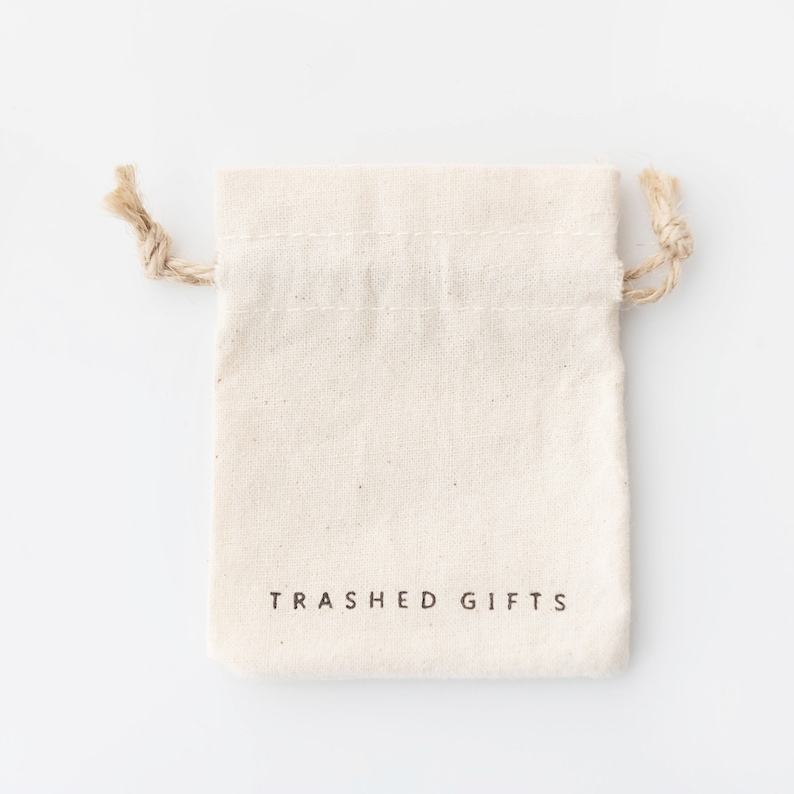 May include: A white cotton drawstring bag with the text "Trashed Gifts" printed on the front.