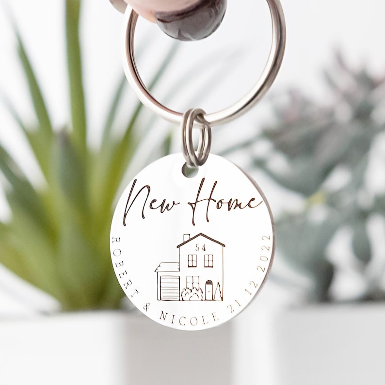 May include: Silver keychain with a round tag that says "New Home" with a house illustration and the names "Robert & Nicole" and the date "21.12.2022".