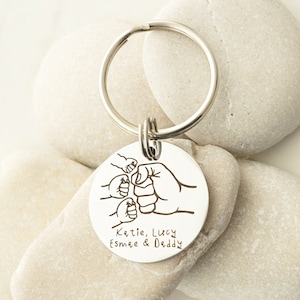 May include: Silver keychain with a round charm featuring four fists bumping together. The charm is engraved with the text "Katie, Lucy, Esmae & Daddy".