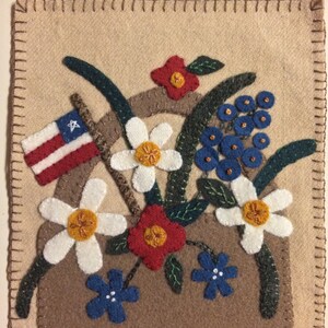May include: A brown felt basket filled with a variety of felt flowers, including white daisies, red and orange flowers, and blue forget-me-nots. An American flag is tucked into the basket.